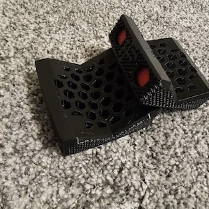 May include: Black 3D-printed phone stand with a honeycomb pattern and two red circular accents. The stand is designed to hold a phone at an angle. The stand is made of plastic.