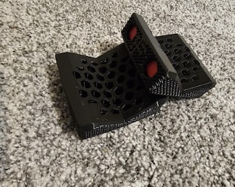 May include: Black 3D-printed phone stand with a honeycomb pattern and two red circular accents. The stand is designed to hold a phone at an angle. The stand is made of plastic.