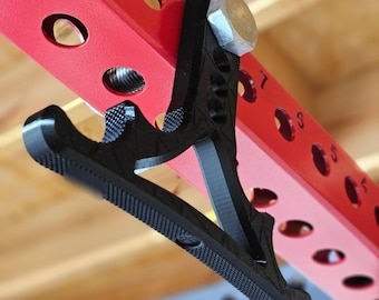 May include: A black metal tool with a textured surface and a hexagonal nut. The tool is attached to a red metal bar with numbered holes.