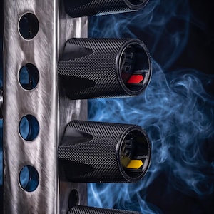 May include: A vertical brushed metal bar with circular cutouts holds four cylindrical black knobs with textured surfaces. Each knob features a small colored indicator inside: blue, red, yellow, and orange, set against a dark background with blue smoke.