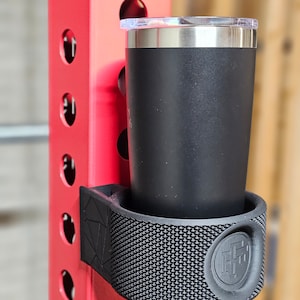 May include: Black metal cup holder with a textured surface and a logo. The holder is attached to a red metal pole with the number "18" on it. A black and silver tumbler is in the holder.