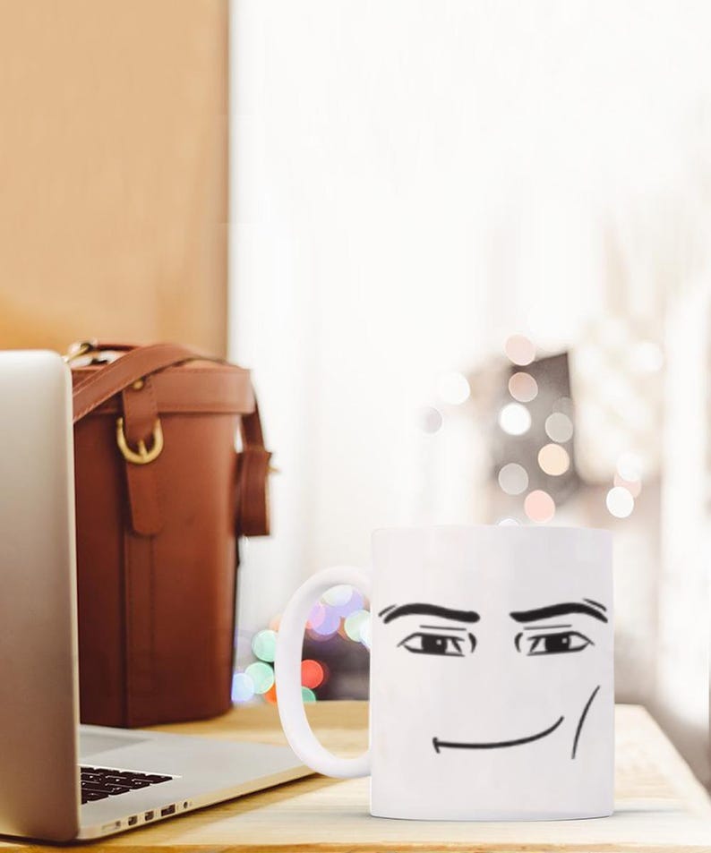 May include: White ceramic coffee mug with a black and white illustration of a face with a smirk. The mug has a handle on the right side.