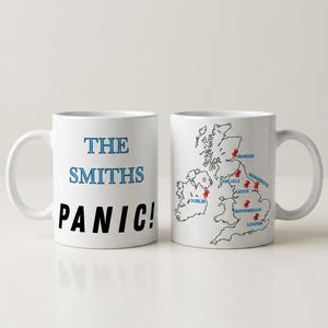 May include: Two white ceramic mugs featuring printed designs. One mug displays the text THE SMITHS in blue and PANIC! in black. The other mug shows a black outline map of the United Kingdom and Ireland with red pin markers on cities including Dublin, Dundee, Carlisle, Leeds, Humberside, Birmingham, and London.
