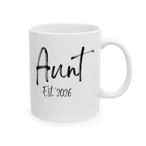 May include: A white ceramic mug featuring the black text Aunt Est. 2026 printed on the front. The mug has a smooth, glossy finish, a cylindrical body, and a curved handle on the right side. The text is written in a combination of brush-style lettering.