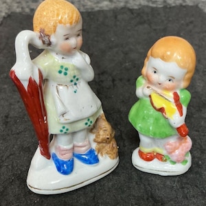 以下が含まれることがあります： 子供の小さな磁器人形が2つ。一人の女の子は赤い傘を持ち、緑と白のドレスに青い靴を履いています。もう一人の女の子は緑のドレスを着て、黄色のバイオリンを持っています。どちらも金髪で頬がバラ色です。