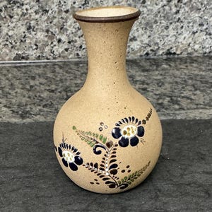 May include: A tan ceramic bud vase with a speckled texture and a narrow neck. The body of the vase is decorated with hand-painted floral motifs in dark blue, green, and white, featuring stylized petals, leaves, and small dots around the base.