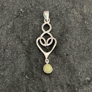 May include: A silver pendant featuring an elongated, openwork design with interlocking loops and a heart-shaped base. A small, round, pale yellow gemstone is suspended from the bottom of the metal structure, which includes a top bail for hanging.