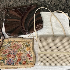 May include: Three handbags: a dark brown leather shoulder bag with a sunburst pattern, a cream-colored tapestry bag with a floral design and metal frame, and a cream-colored metal mesh shoulder bag with a rectangular flap and thin strap.