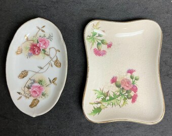May include: Two decorative porcelain dishes. One is oval-shaped with pink roses and gold accents. The other is a rectangular dish with a curved edge, featuring pink thistle flowers and gold trim. Both have a cream-colored background.