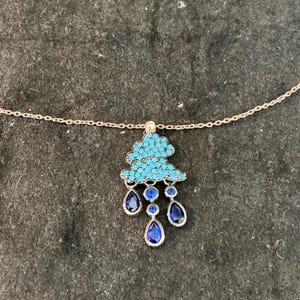 May include: A rose gold chain necklace featuring a cloud-shaped pendant encrusted with small turquoise stones. Three dangling blue teardrop-shaped crystals and two small round blue crystals hang from the bottom of the cloud to resemble rain.