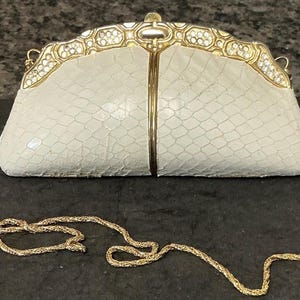 May include: A cream-colored clutch bag with a textured snakeskin-like pattern, featuring a gold-tone metal frame adorned with clear crystals along the top edge, a central vertical gold bar, and a detachable thin gold-tone metal chain shoulder strap.