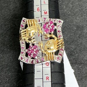 May include: A gold and silver ring featuring two crab designs with ruby accents. The ring has a rectangular shape with a silver border and is set on a black ring sizer. The crabs are gold, and the rubies are a deep pink.