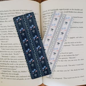 May include: Two patterned bookmarks, one with a dark blue background and the other with a white background. Both bookmarks feature a repeating pattern of blue and pink flowers with green leaves and white bows.