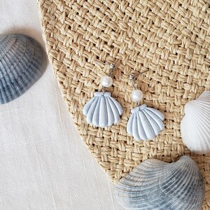 May include: A pair of light blue seashell earrings with a pearl accent. The earrings are on a woven straw background.
