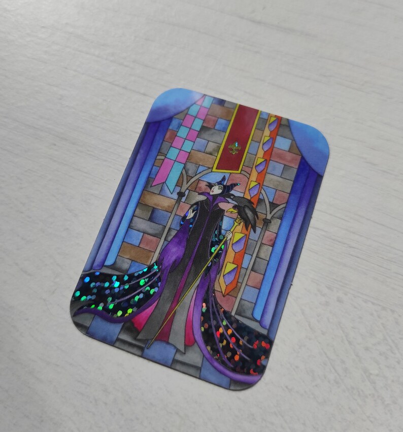 K&ouml;nnte beinhalten: Ein Aufkleber mit einer Cartoon-Illustration von Maleficent aus Disneys Dornr&ouml;schen. Sie steht in einem Buntglasfenster mit ihrem Raben und h&auml;lt einen Stab. Der Aufkleber hat ein glitzerndes lila und blaues Design.