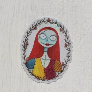 K&ouml;nnte beinhalten: Ein runder Aufkleber mit wei&szlig;em Hintergrund und glitzerndem Finish. Der Aufkleber zeigt eine Cartoon-Illustration von Sally aus Nightmare Before Christmas. Sie hat rote Haare, blaue Haut und tr&auml;gt ein buntes Patchwork-Kleid. Sie ist von einem Kranz aus braunen Zweigen mit roten Bl&uuml;ten umgeben.