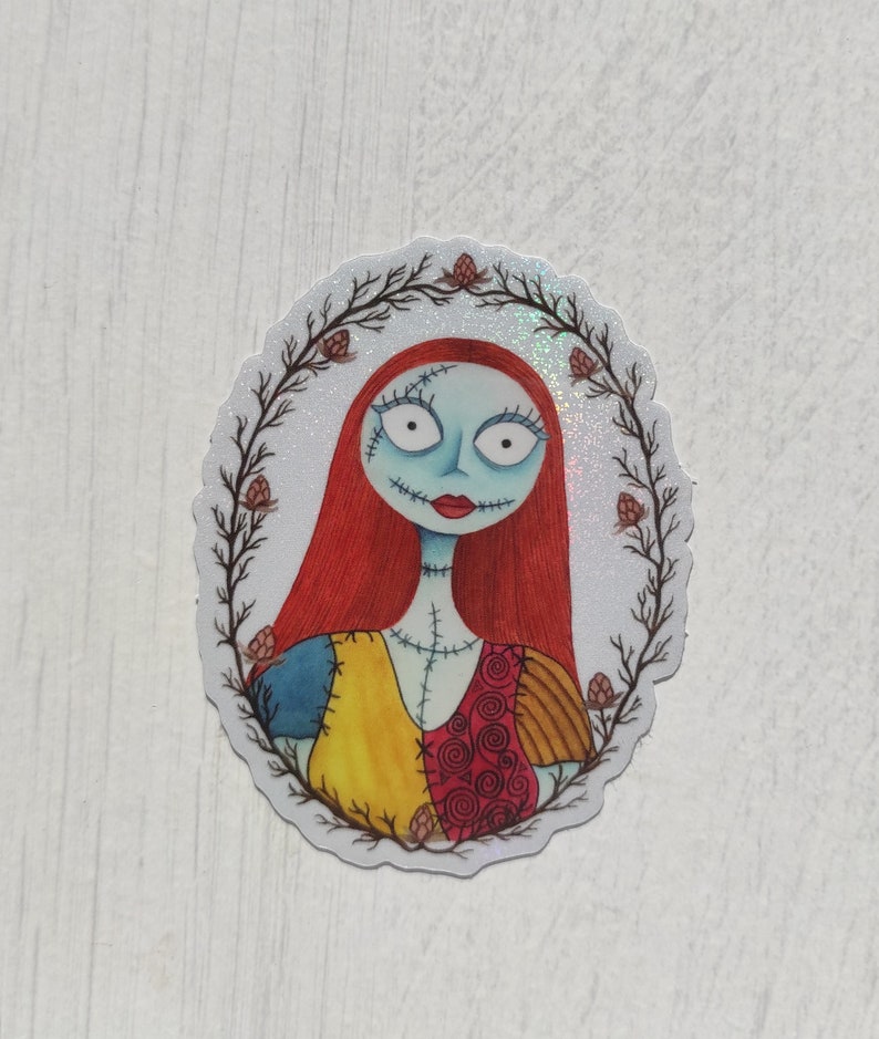 K&ouml;nnte beinhalten: Ein runder Aufkleber mit wei&szlig;em Hintergrund und glitzerndem Finish. Der Aufkleber zeigt eine Cartoon-Illustration von Sally aus Nightmare Before Christmas. Sie hat rote Haare, blaue Haut und tr&auml;gt ein buntes Patchwork-Kleid. Sie ist von einem Kranz aus braunen Zweigen mit roten Bl&uuml;ten umgeben.