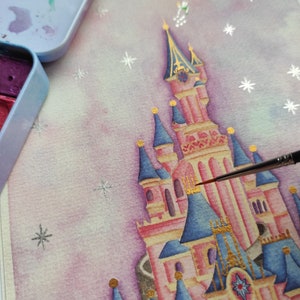 Op de afbeelding: Een aquarel schilderij van een roze en blauw kasteel met gouden accenten. Het kasteel staat tegen een lichtpaarse en witte achtergrond met zilveren glittersterren.