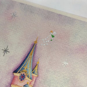 Op de afbeelding: Een roze en blauw kasteel met gouden accenten tegen een paarse en witte aquarel achtergrond. Een kleine groene fee met een gouden kroon vliegt boven het kasteel en strooit zilveren glitter.