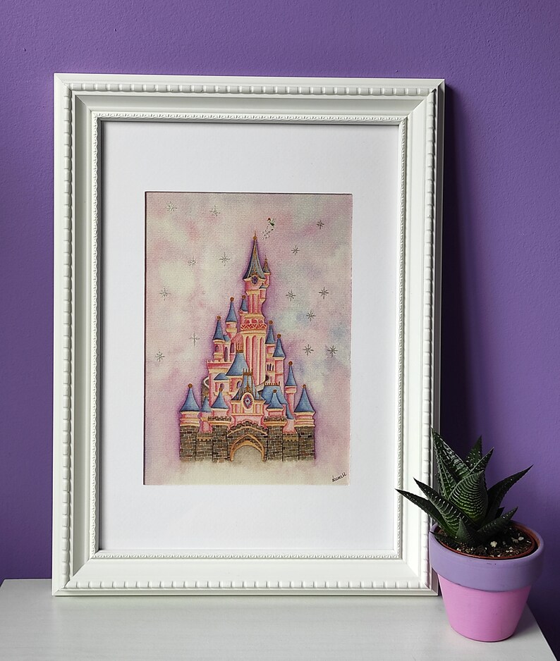 Op de afbeelding: Een ingelijst aquarel schilderij van een roze kasteel met blauwe torens en een witte achtergrond met sterren. Het kasteel is omgeven door een licht roze en blauwe lucht. Het schilderij is in een witte lijst.