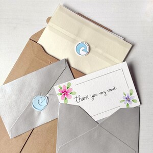 Op de afbeelding: Een bedankkaartje met een handgeschreven bericht "Hartelijk dank" en aquarelbloemen in roze en paars. Het kaartje zit in een grijze envelop met een blauwe waszegel.