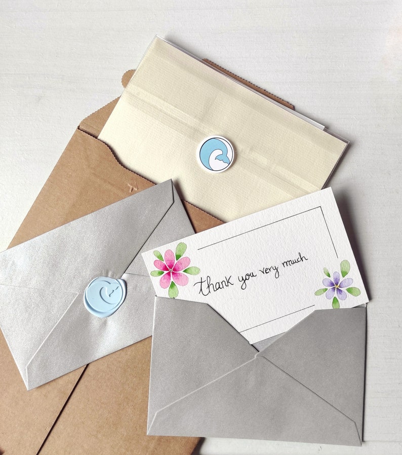Op de afbeelding: Een bedankkaartje met een handgeschreven bericht "Hartelijk dank" en aquarelbloemen in roze en paars. Het kaartje zit in een grijze envelop met een blauwe waszegel.