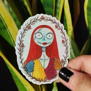 K&ouml;nnte beinhalten: Ein wei&szlig;er Aufkleber mit glitzernder Oberfl&auml;che, der eine Cartoon-Illustration von Sally aus Nightmare Before Christmas zeigt. Sally tr&auml;gt ihr charakteristisches Patchwork-Kleid und hat rote Haare. Der Aufkleber ist von einem Kranz aus braunen Zweigen mit roten Beeren umgeben.