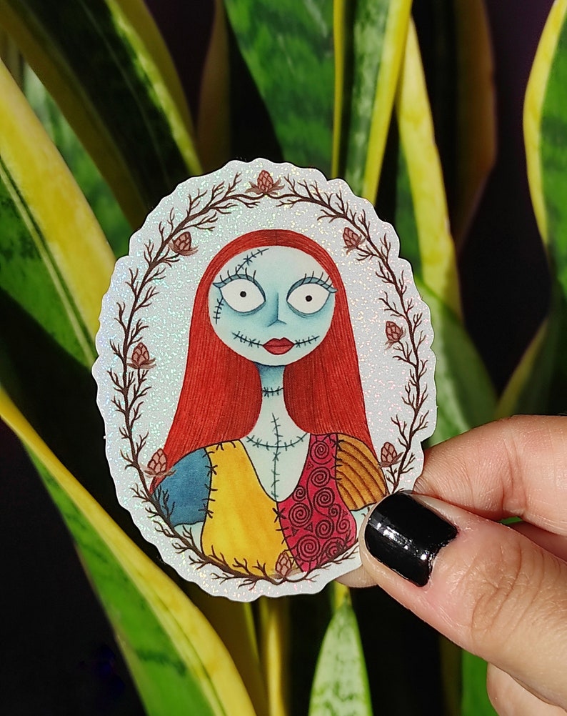 K&ouml;nnte beinhalten: Ein wei&szlig;er Aufkleber mit glitzernder Oberfl&auml;che, der eine Cartoon-Illustration von Sally aus Nightmare Before Christmas zeigt. Sally tr&auml;gt ihr charakteristisches Patchwork-Kleid und hat rote Haare. Der Aufkleber ist von einem Kranz aus braunen Zweigen mit roten Beeren umgeben.