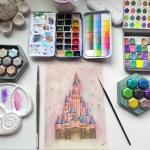Op de afbeelding: Een aquarel schilderij van Assepoester's kasteel met een roze en blauwe kleurstelling. Het kasteel is omringd door een lichtroze achtergrond met witte sterren. Het schilderij is op een witte passe-partout en is ingelijst met een zwarte lijst.