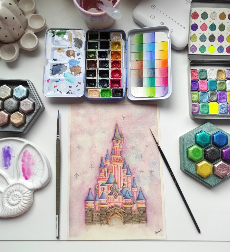 Op de afbeelding: Een aquarel schilderij van Assepoester's kasteel met een roze en blauwe kleurstelling. Het kasteel is omringd door een lichtroze achtergrond met witte sterren. Het schilderij is op een witte passe-partout en is ingelijst met een zwarte lijst.