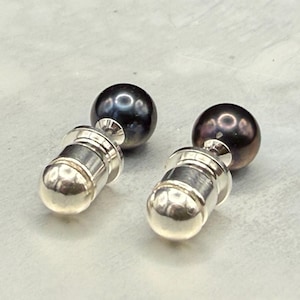May include: A pair of cufflinks featuring dark, lustrous pearls atop silver-colored cylindrical bases. The pearls have a deep, iridescent sheen, reflecting light. The cufflinks are presented on a light gray surface.