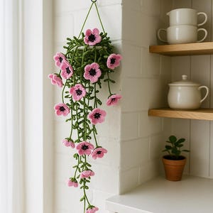May include: A handmade crochet hanging basket with pink flowers and green leaves. The flowers have dark centers and are arranged in a cascading pattern. The basket is suspended from a black hook on a white brick wall, adding a touch of handmade charm to any space.