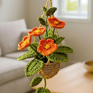 May include: A handmade crochet hanging planter with orange flowers and green leaves. The flowers have yellow centers. The planter is suspended by a yellow cord and is in a beige pot. The background is blurred, showing a living room setting.