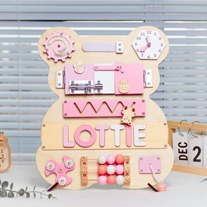 May include: A wooden activity board shaped like a bear with a pink and white color scheme. The board features a variety of activities for children, including a clock, a latch, a sliding door, a shape sorter, a bead maze, a spinning wheel, and a name puzzle that reads "LOTTIE".