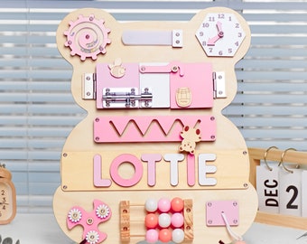 Puede incluir: Un tablero de actividades de madera con forma de oso con un esquema de color rosa y blanco. El tablero presenta una variedad de actividades para niños, incluyendo un reloj, un pestillo, una puerta corredera, un clasificador de formas, un laberinto de cuentas, una rueda giratoria y un rompecabezas de nombre que dice "LOTTIE".