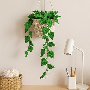 May include: A handmade crochet hanging planter with a woven basket design, filled with vibrant green leaves. The plant hangs from a wooden wall mount, adding a touch of nature to any space. The leaves cascade downwards, creating a visually appealing display.