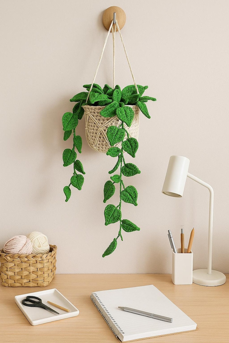 May include: A handmade crochet hanging planter with a woven basket design, filled with vibrant green leaves. The plant hangs from a wooden wall mount, adding a touch of nature to any space. The leaves cascade downwards, creating a visually appealing display.