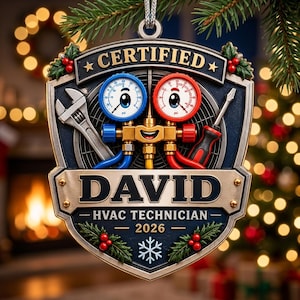 Op de afbeelding: Een schildvormig kerstornament met een lachend HVAC-manometerfiguurtje, een moersleutel en een schroevendraaier. De tekst luidt CERTIFIED, DAVID, HVAC TECHNICIAN en 2026, versierd met hulstbessen en een sneeuwvlokje in reliëf.