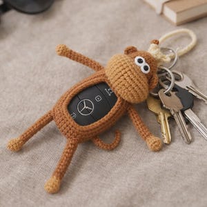 Può includere: Una custodia per chiavi all'uncinetto marrone a forma di scimmia, con testa rotonda, due occhi in plastica bianca e nera, quattro arti e una piccola coda. Il corpo della scimmia avvolge una chiave auto Mercedes-Benz nera, con un anello metallico.