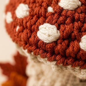 May include: A crocheted mushroom figure featuring a deep rust-colored cap adorned with raised, off-white circular spots. The stem is crafted from a light cream-colored yarn with a textured, ribbed appearance, showcasing detailed hand-stitched fiber work.