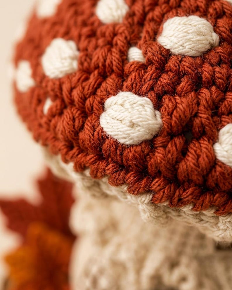 May include: A crocheted mushroom figure featuring a deep rust-colored cap adorned with raised, off-white circular spots. The stem is crafted from a light cream-colored yarn with a textured, ribbed appearance, showcasing detailed hand-stitched fiber work.