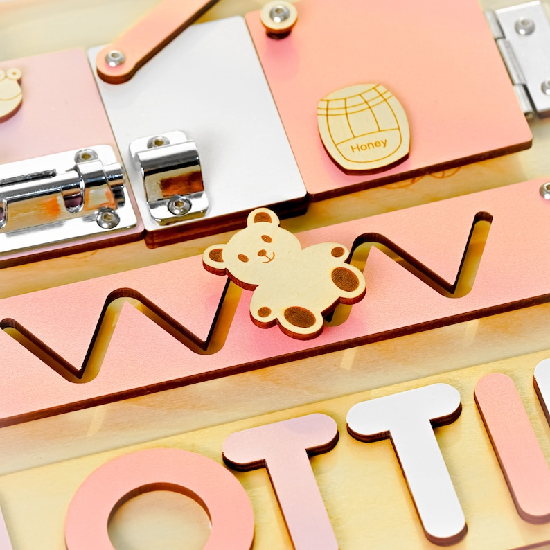 May include: A pink and yellow wooden busy board with a variety of activities for children. The board features a teddy bear, a latch, a sliding door, a maze, and the word "LOT" in large letters.