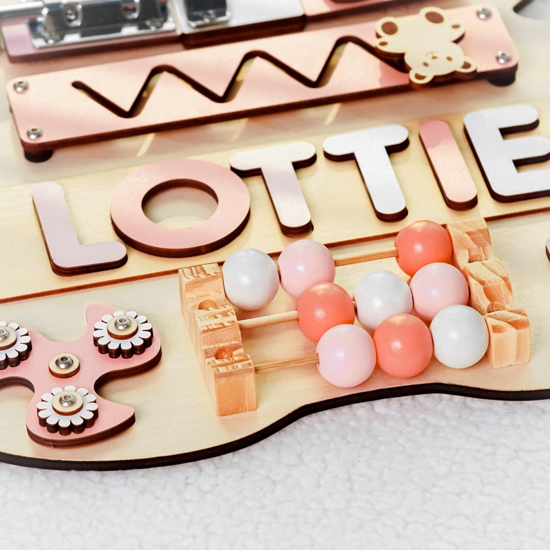 May include: A wooden activity board with the name "Lottie" spelled out in pink letters. The board features a variety of activities for children, including a bead maze, a spinning fidget toy, and a shape sorter.
