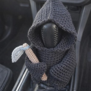 以下が含まれることがあります： 手作りの車のアクセサリー、ダークグレーのかぎ針編みの死神。フードと小さなつるはしが付いています。車のギアシフトにフィットするように設計されており、車のインテリアにユニークなタッチを加えます。