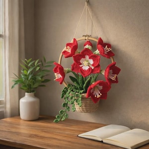 Puede incluir: Un colgante de pared floral de ganchillo hecho a mano con flores rojas vibrantes con centros blancos y amarillos, dispuestas en un aro de bordado de madera circular. Las flores están colocadas en una cesta tejida con follaje verde, suspendida por hilo natural.