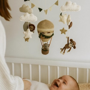 May include: A handmade wooden baby mobile featuring crocheted clouds, stars, and a hot air balloon with a monkey and lion. The mobile is suspended above a baby in a crib. The mobile is made of beige, brown, and white yarn.
