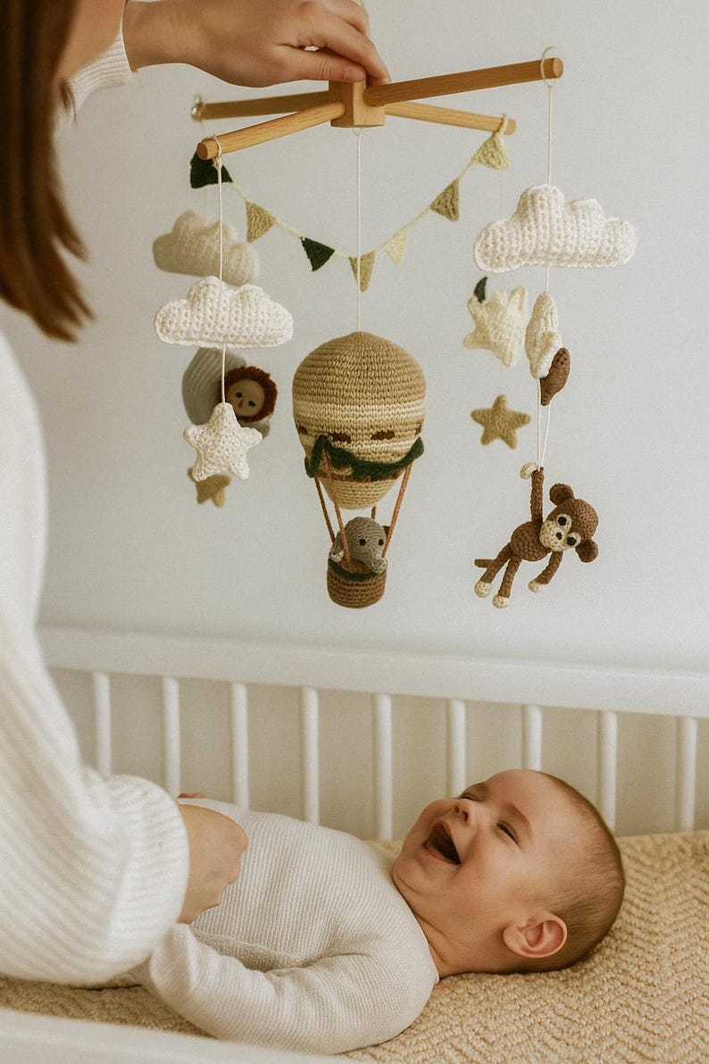 May include: A handmade wooden baby mobile featuring crocheted clouds, stars, and a hot air balloon with a monkey and lion. The mobile is suspended above a baby in a crib. The mobile is made of beige, brown, and white yarn.