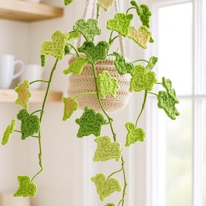 May include: A handmade crochet hanging plant with green ivy leaves. The plant is in a beige crochet pot suspended by a cream-colored rope. The leaves vary in shades of green and yellow.