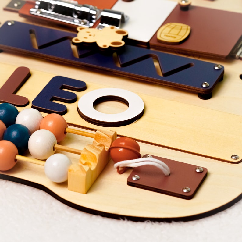 May include: A wooden activity board with a blue and white nameplate that reads "LEO". The board features a variety of textures and activities for children, including a bead maze, a sliding door, and a shape sorter.