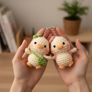 Könnte beinhalten: Zwei handgefertigte Häkel-Enten-Schlüsselanhänger. Eine Ente hat eine grüne Schleife und Tasche, die andere eine rosa Schleife. Beide haben orangefarbene Schnäbel und Füße sowie schwarze Augen. Die Schlüsselanhänger sind durch eine silberne Kette verbunden.